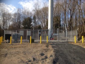 Here is a steel chainlink fence equipped with barbed wire, with cement bollards placed in front of it. 